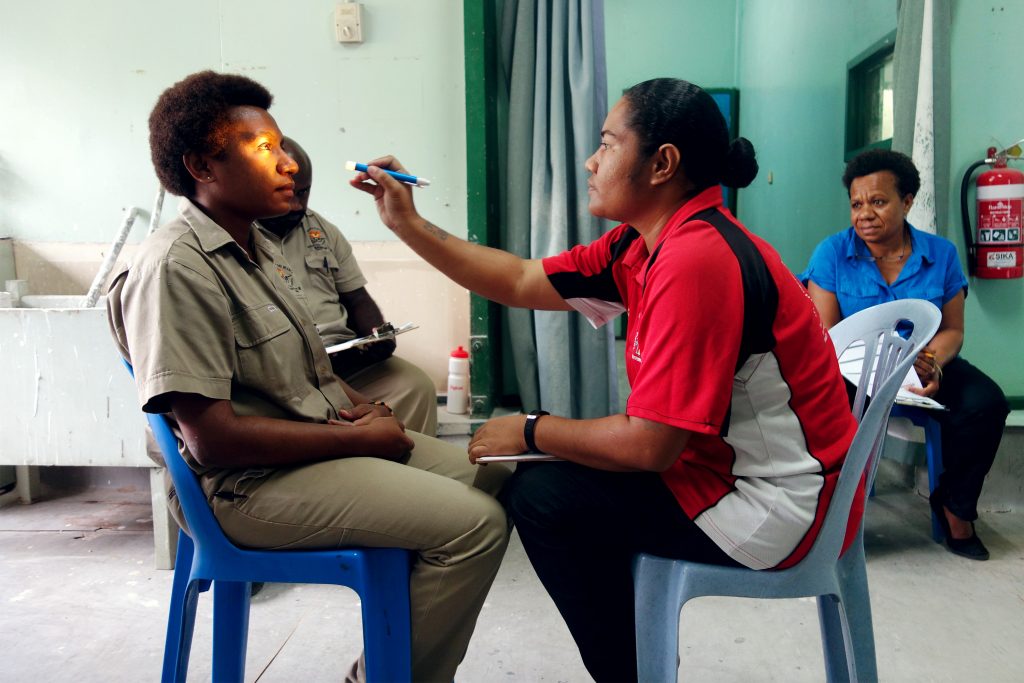 A service provider with a clipboard uses a torch to check the eyes of another woman.