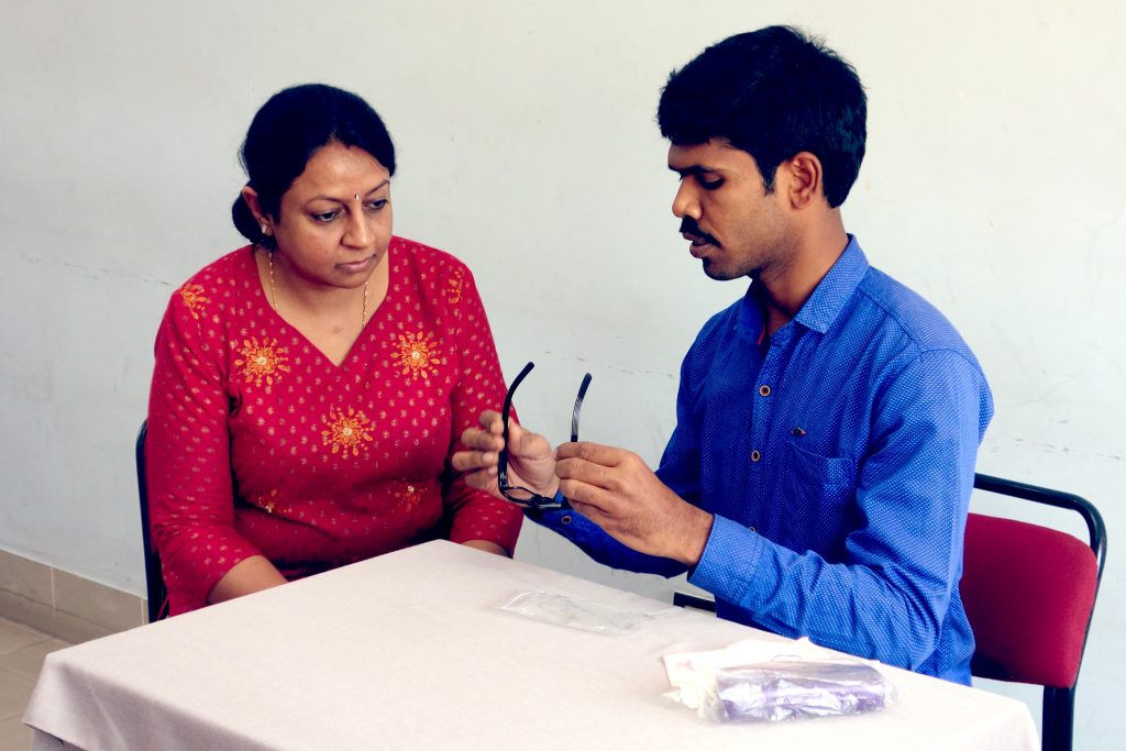 Two people sitting at a table. A man is holding glasses and showing them to the woman.