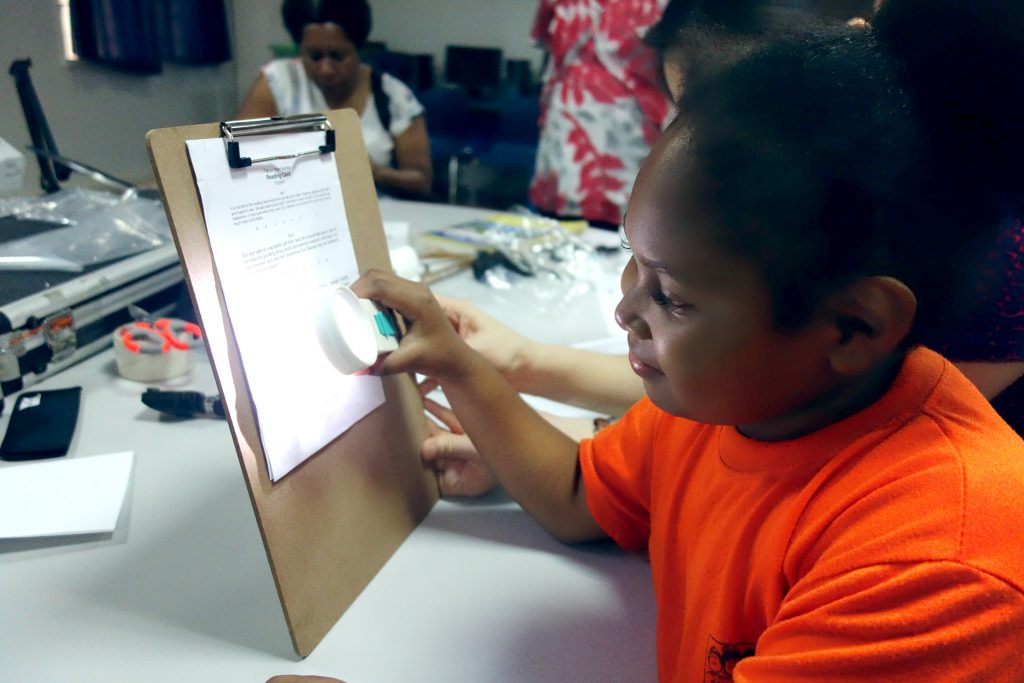 A young girl smiles as she uses a magnifying glass with a light to read text presented to her on a clipboard.