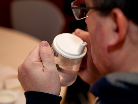 A man wearing glasses uses a modified cup with handles and a lid to drink.