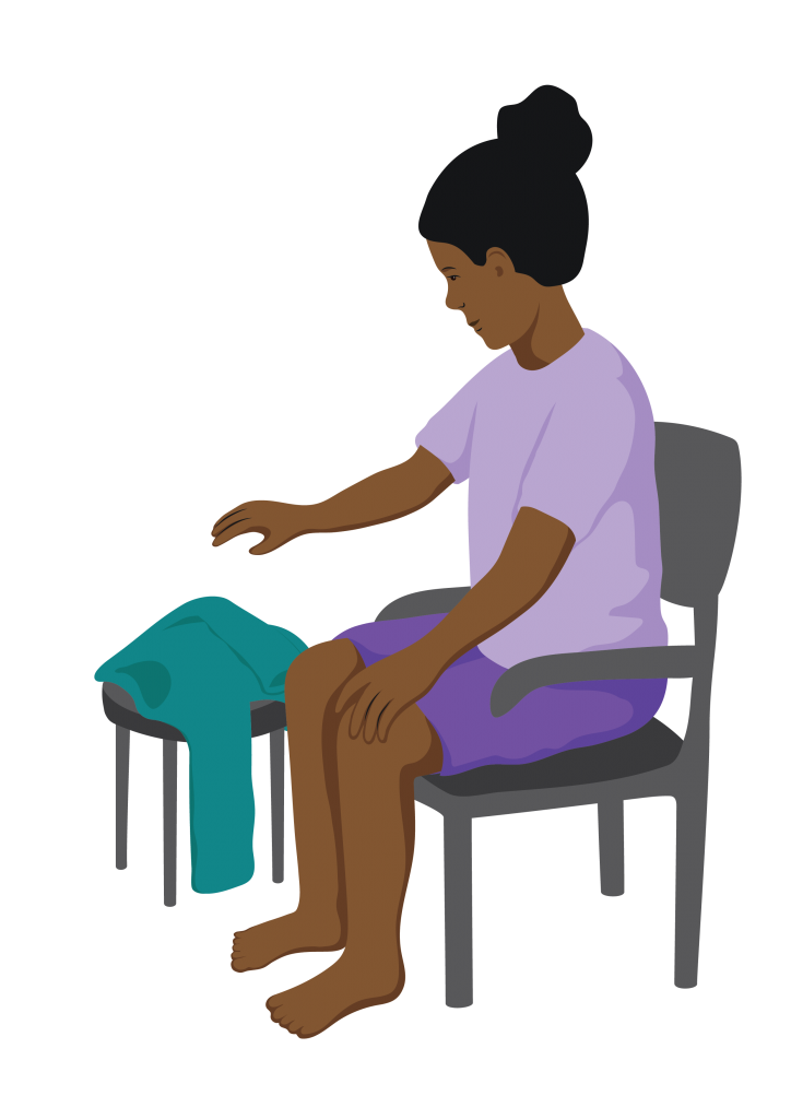 Aleisha is sitting on a chair with arm rests and picks up a jumper from a stool nearby.