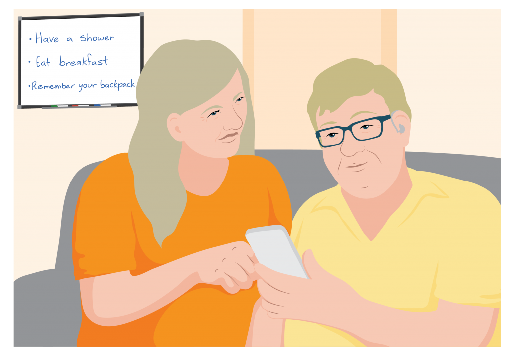 Samuel holds a mobile phone. A woman sitting next to him points at the device and explains how to use a function. A whiteboard with reminders written on it is in the background.