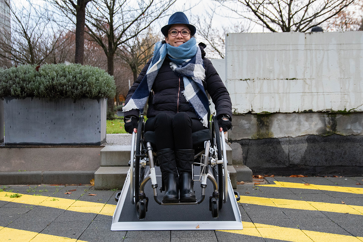 A woman dressed warmly in a coat and scarf uses a wheelchair and portable ramp to go down two steps.