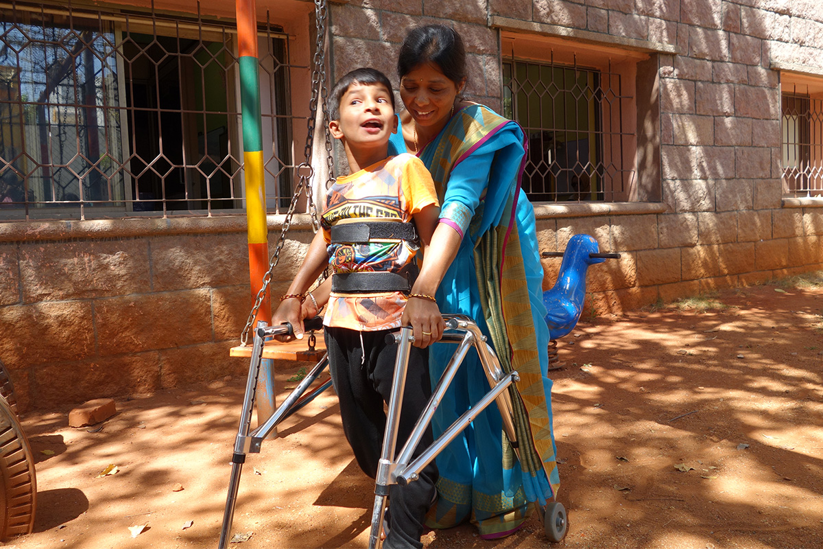 A woman stands behind a boy who is using a posterior walker. They are both holding onto the frame. The boy is wearing a back brace. Together they are walking in the shade of a tree.