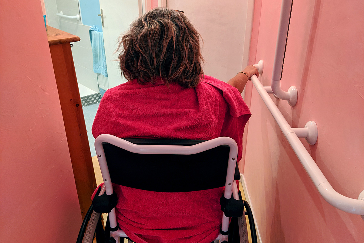 A woman in a wheelchair uses grab bars to help her move down a hallway towards a bathroom.