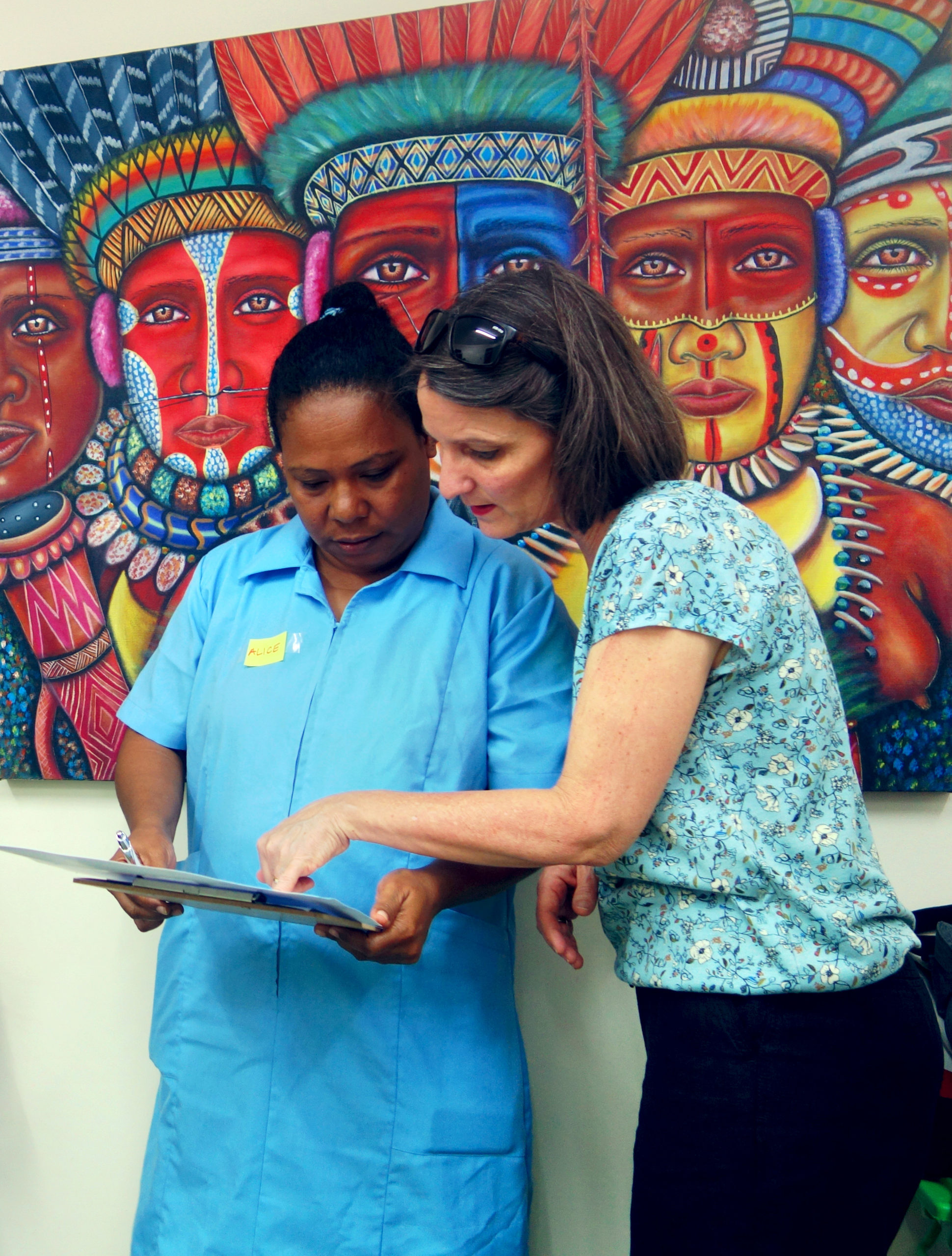 Two women stand in front of a colourful painting of people in traditional papua new guinea dress. One woman is holding a clipboard, another woman is leaning over to point at something on the clipboard.