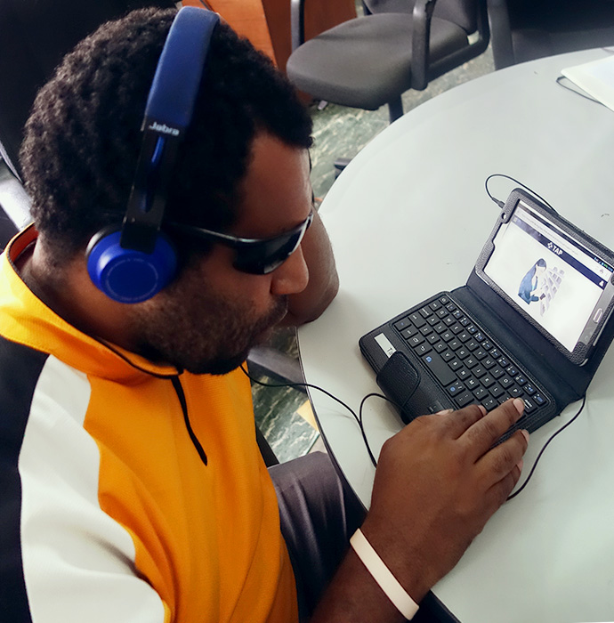 A man wearing dark glasses and headphones uses a tablet connected to a keyboard to use TAP.