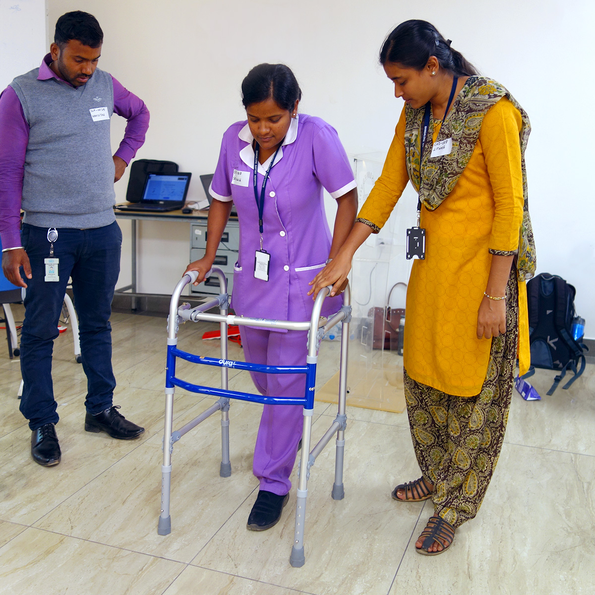 A woman in a service uniform uses a walking frame. Another woman has her hand on the frame for support and is watching closely. A man stands to the side also watching.