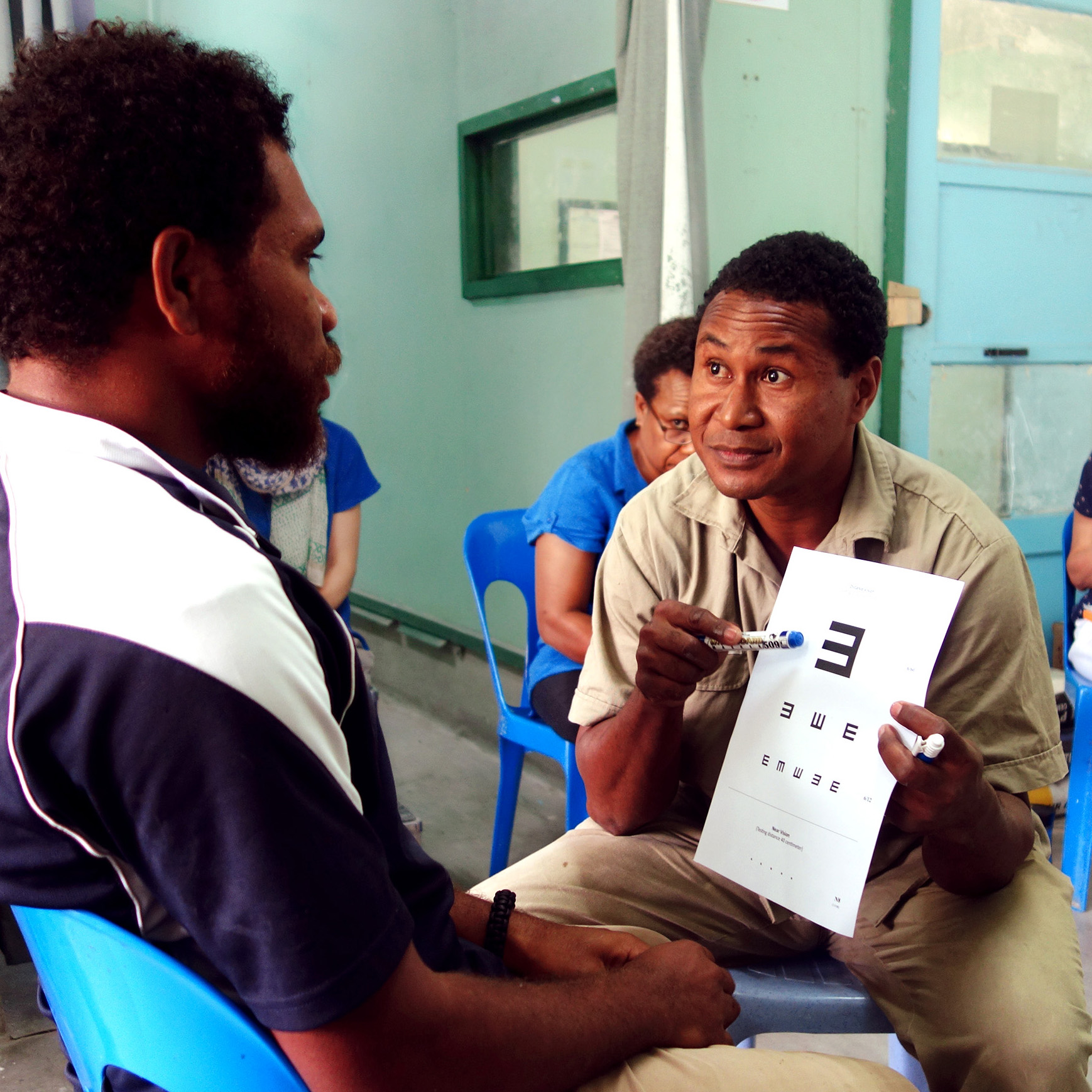 Two men are sitting down. The man on the right is holding up a vision screen E chart and gesturing to the large E with a pen. The man on the left is looking at the vision chart.