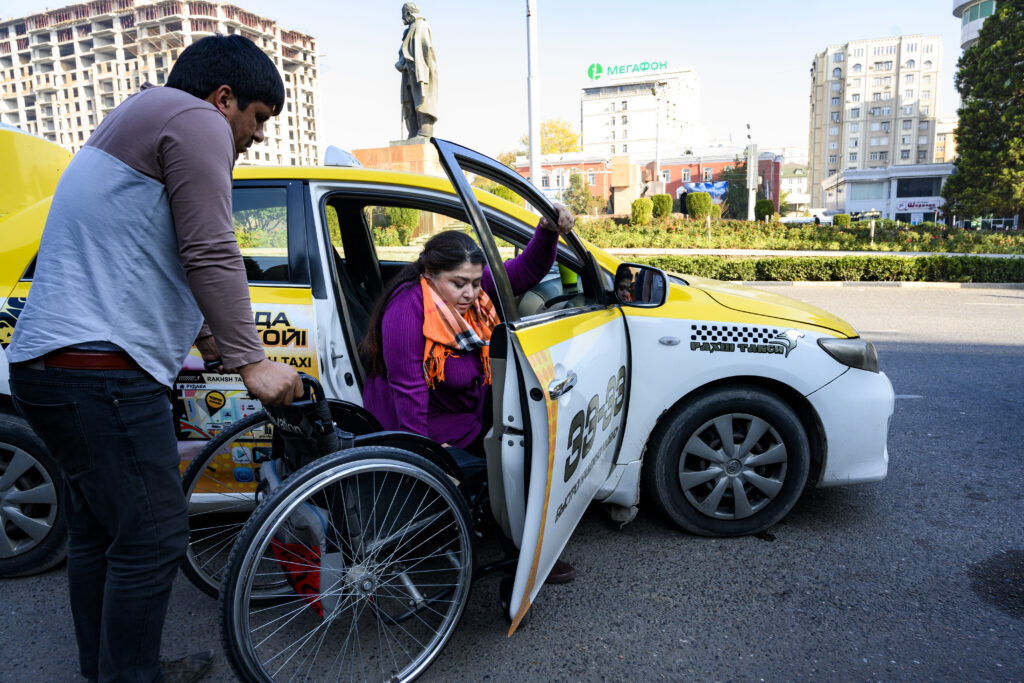 A woman transfers from a car to a wheelchair. A man assists by holding onto the push handles of the wheelchair.