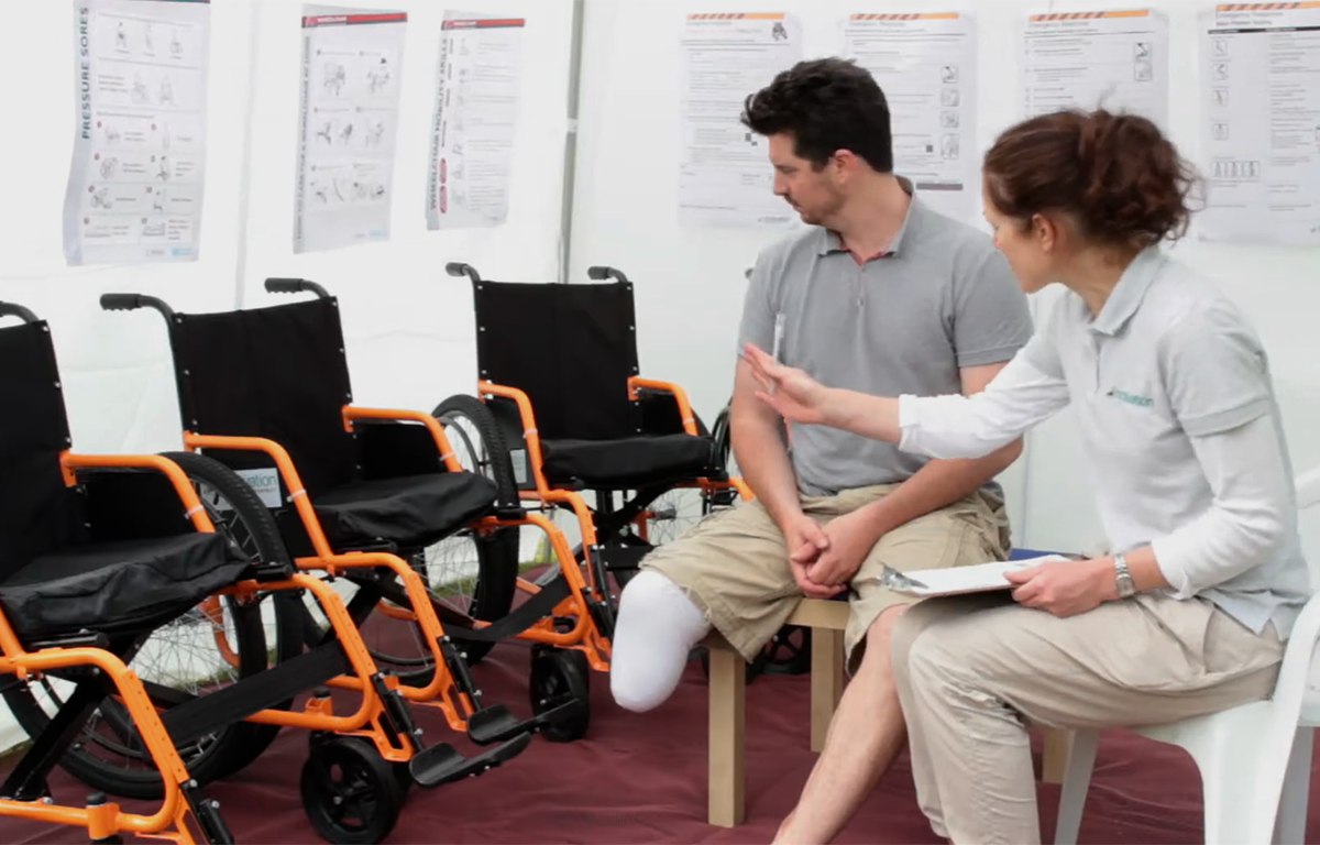 A man with a below knee amputation and a female health worker look over at a row of three different size wheelchairs.