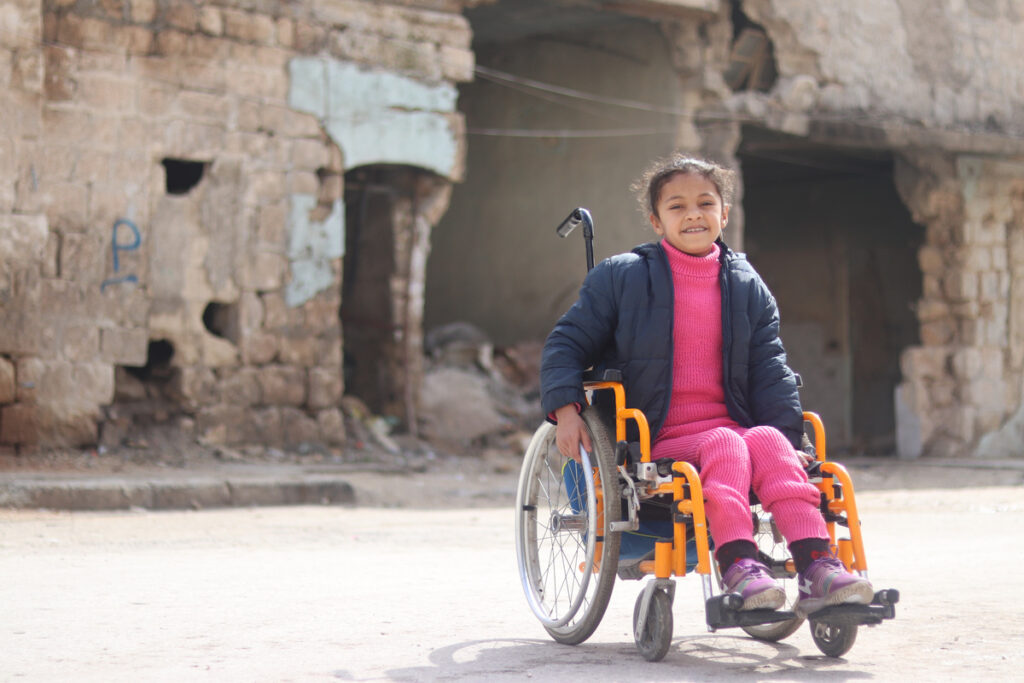 A young girl sits in a wheelchair, smiling at the camera. Behind her are damaged houses.