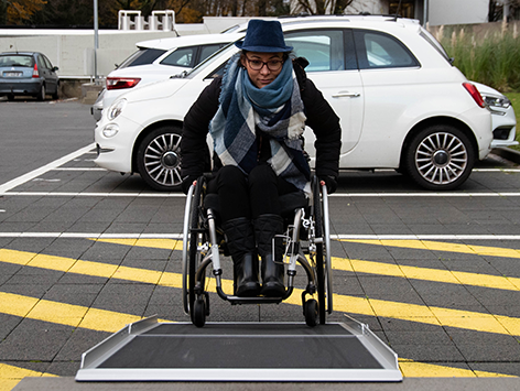 Woman using a four-wheel wheelchair pushing up a wide platform portable ramp in a car park.