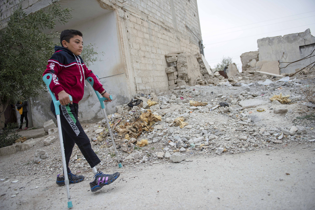 A child walking with elbow crutches. He has prosthetic legs. There is a collapsed building behind the child.