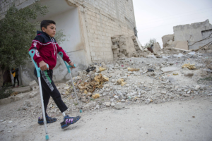 A child walking with elbow crutches. He has prosthetic legs. There is a collapsed building behind the child.