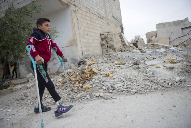 A child walking with elbow crutches. He has prosthetic legs. There is a collapsed building behind the child.