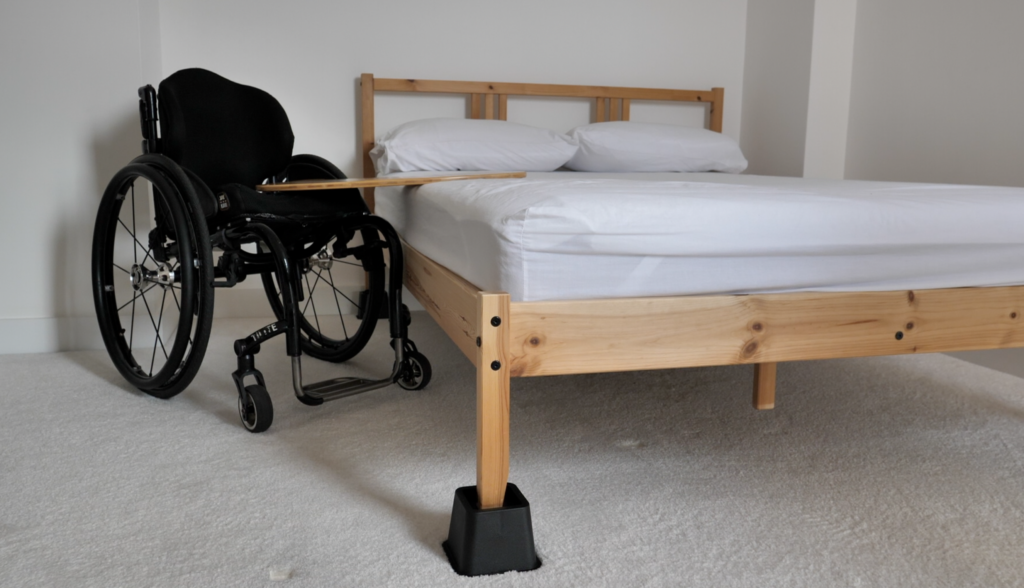 One end of a curved wooden transfer board is placed on a wheelchair and the other end on a bed. The bed has bed raisers to create a similar height between the wheelchair and the bed.
