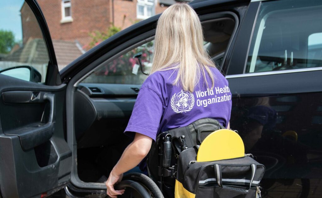 A woman sitting in a wheelchair with a backpack hanging over the backrest. The backpack holds a transfer board. The person is in front of a car.