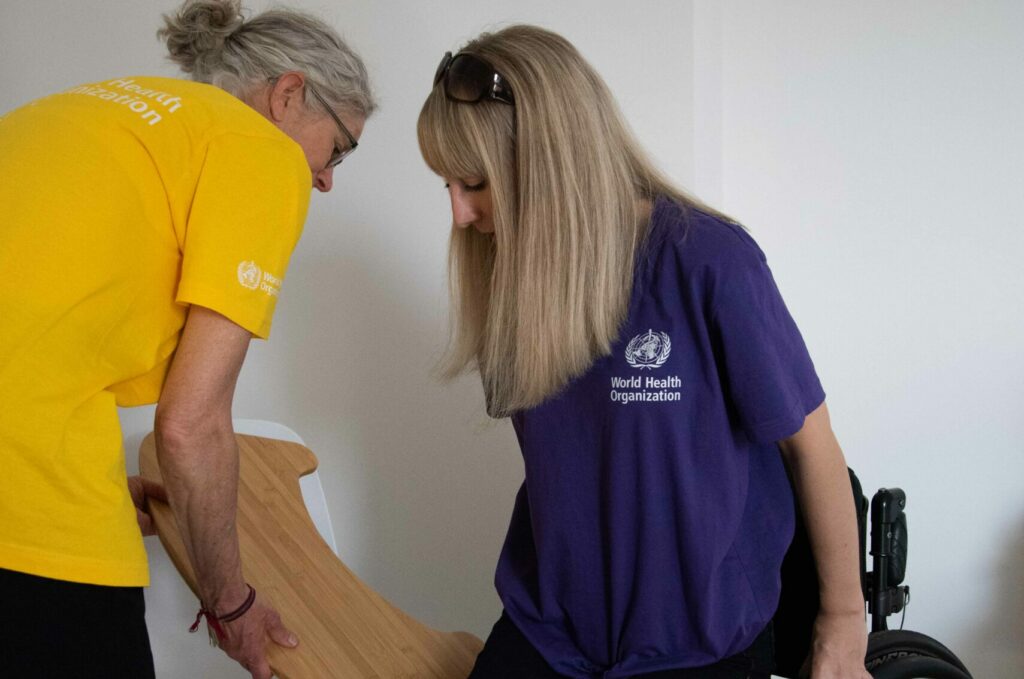 A woman in a wheelchair is lifting up off their wheelchair to start a wheelchair to chair transfer. A health worker is holding a curved transfer board.