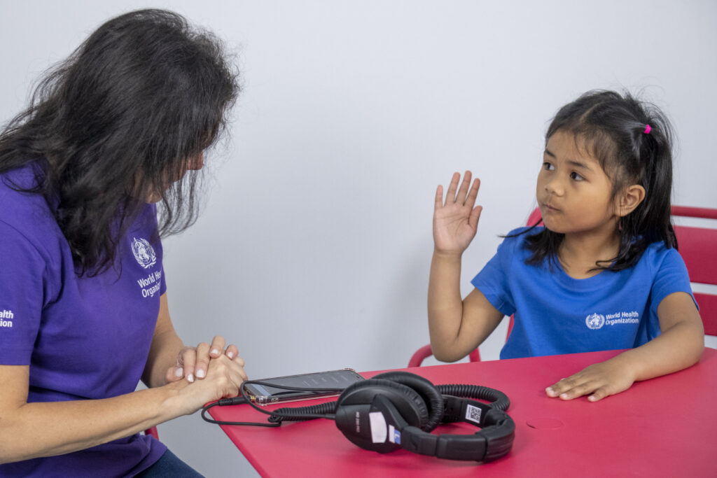 A health worker speaking to a child. There is a phone connected to headphones on the table. The child is raising their right hand in a practice test response.