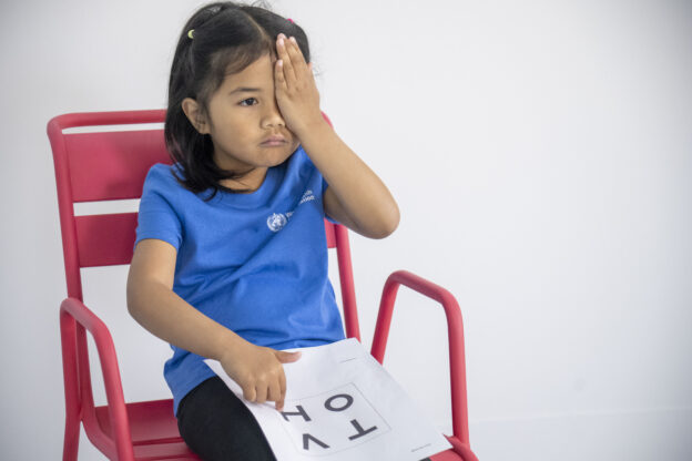 Child sitting on chair. One hand points to letter on pointing chart and other hand covers one eye.