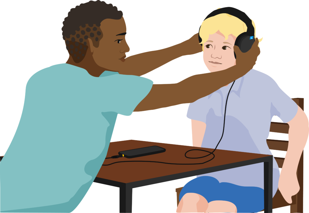 The screener and child are sitting and leaning towards each other across a table. The screener places headphones over the child’s ears.