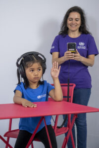 A child sits at a table with their left hand raised. The chid wears headphones. The headphones are connected to a phone. The phone is in the hands of a health worker who is standing behind the child.